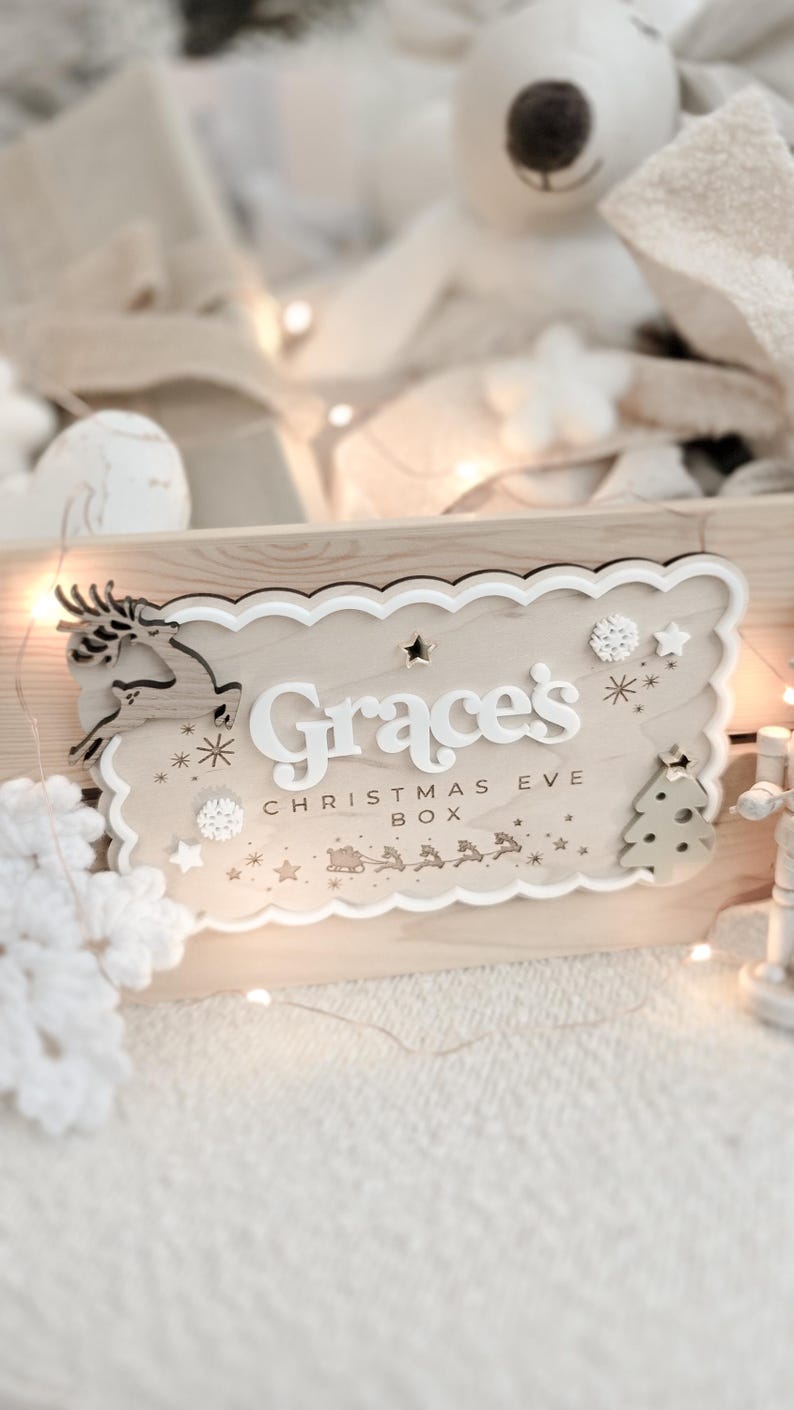 May include: A wooden Christmas Eve box with a scalloped edge, featuring the name "Grace's" in white letters. The box is decorated with a reindeer, stars, snowflakes, and a Christmas tree. The words "CHRISTMAS EVE BOX" are also visible.