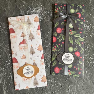 Könnte beinhalten: Zwei Geschenkverpackungen mit Weihnachtsmotiven. Eine zeigt Weihnachtsmann-Gesichter und Weihnachtsbäume auf weißem Grund, gebunden mit einem Band. Die andere hat einen schwarzen Hintergrund mit Kugeln und Stechpalmen, gebunden mit einem grauen Band. Beide haben ein Etikett mit "Frohe Weihnachten".