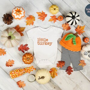 May include: A white onesie with the text "little turkey" in orange. The onesie is surrounded by fall-themed decorations, including pumpkins, leaves, and candy corn.
