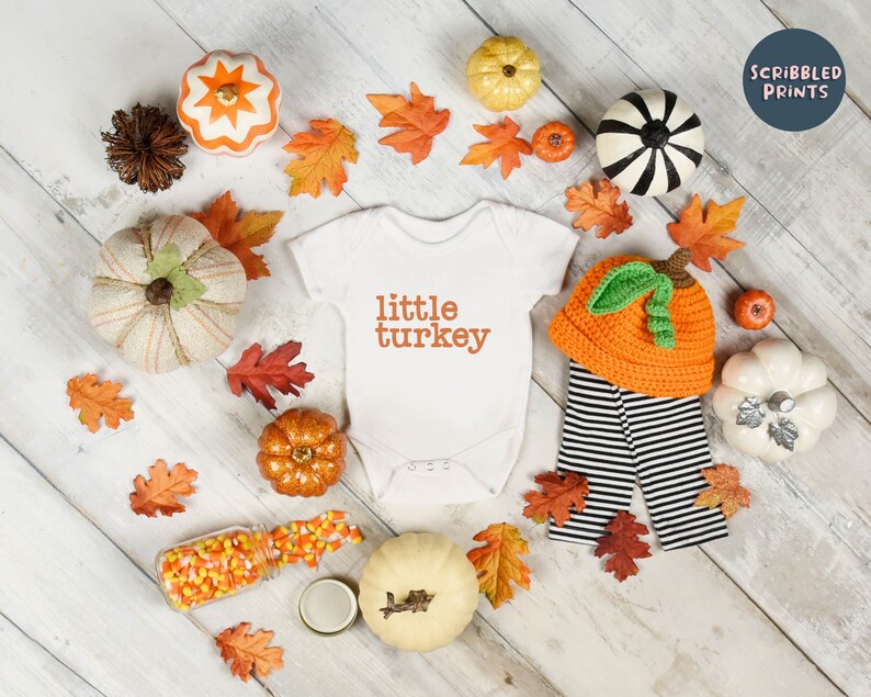 May include: A white onesie with the text "little turkey" in orange. The onesie is surrounded by fall-themed decorations, including pumpkins, leaves, and candy corn.