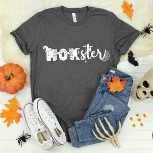 K&ouml;nnte beinhalten: Ein graues T-Shirt mit dem Text "Momster" in Wei&szlig;, verziert mit Halloween-Motiven. Das Shirt liegt auf einer wei&szlig;en Oberfl&auml;che mit Herbstbl&auml;ttern, einem K&uuml;rbis, Maisbonbons, einer Spinne, einer Fledermaus, einem Sch&auml;del und einem Paar wei&szlig;en Sneakers.