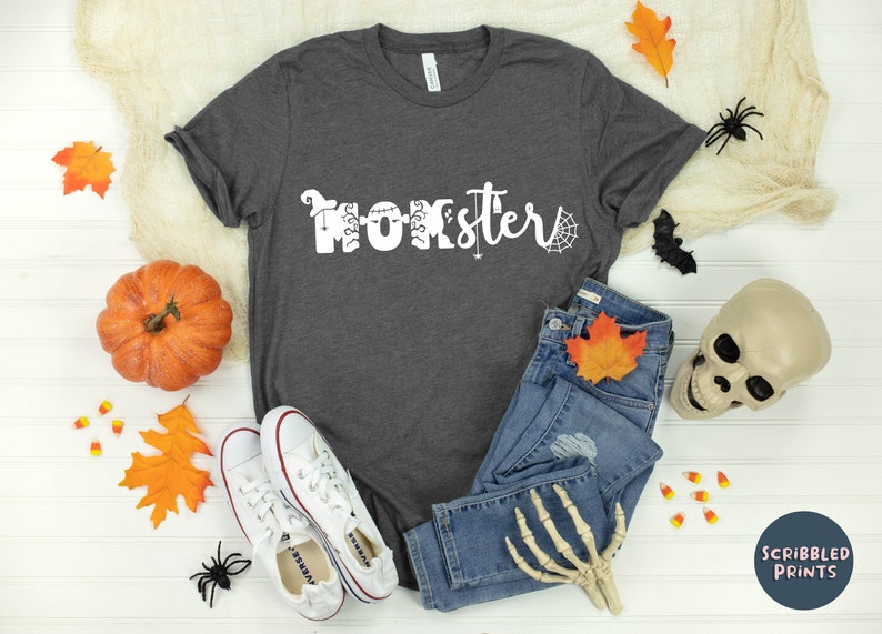 K&ouml;nnte beinhalten: Ein graues T-Shirt mit dem Text "Momster" in Wei&szlig;, verziert mit Halloween-Motiven. Das Shirt liegt auf einer wei&szlig;en Oberfl&auml;che mit Herbstbl&auml;ttern, einem K&uuml;rbis, Maisbonbons, einer Spinne, einer Fledermaus, einem Sch&auml;del und einem Paar wei&szlig;en Sneakers.