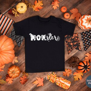 K&ouml;nnte beinhalten: Schwarzes T-Shirt mit wei&szlig;em Text, der "Momster" mit einem Spinnennetz-Design sagt. Das Shirt ist umgeben von Herbstbl&auml;ttern, K&uuml;rbissen und Halloween-Dekorationen.