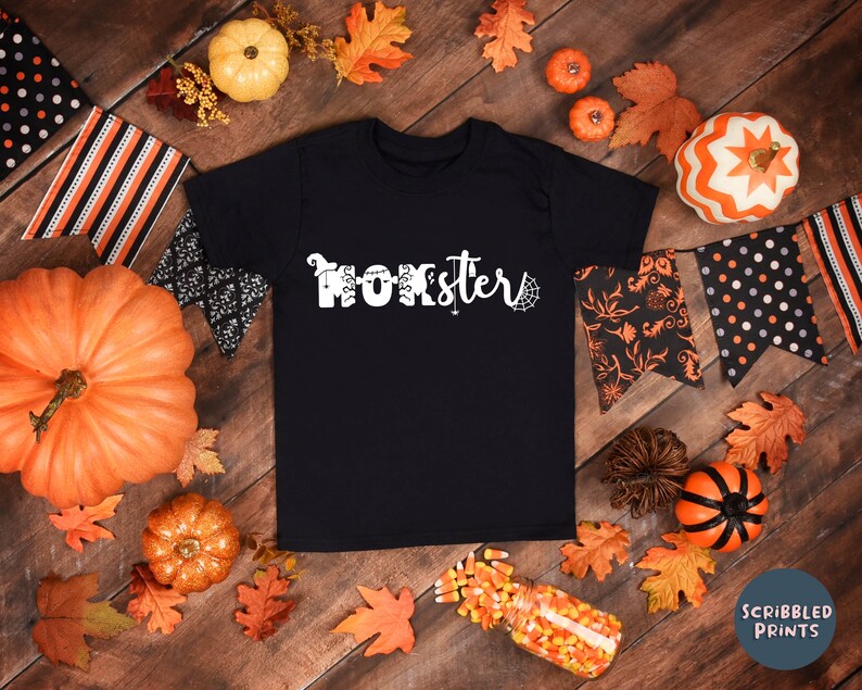K&ouml;nnte beinhalten: Schwarzes T-Shirt mit wei&szlig;em Text, der "Momster" mit einem Spinnennetz-Design sagt. Das Shirt ist umgeben von Herbstbl&auml;ttern, K&uuml;rbissen und Halloween-Dekorationen.