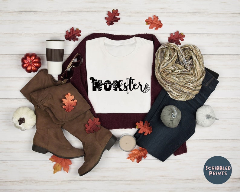 K&ouml;nnte beinhalten: Wei&szlig;es T-Shirt mit dem Text "Monster Sister" in schwarzen Buchstaben mit einer Spinne und einem Geister-Motiv. Das Shirt liegt auf einer wei&szlig;en Holzoberfl&auml;che mit Herbstdeko, darunter braune Stiefel, ein Schal, Jeans und Herbstbl&auml;tter.