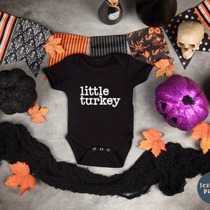 May include: Black baby onesie with the text "little turkey" in white. The onesie is surrounded by Halloween decorations, including a black and orange banner, a purple glitter pumpkin, and a black skull.