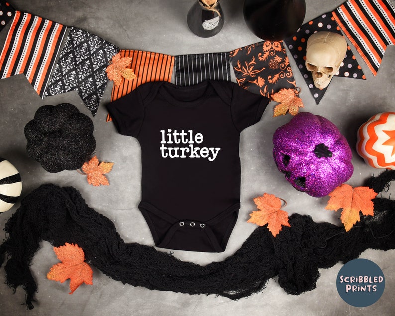 May include: Black baby onesie with the text "little turkey" in white. The onesie is surrounded by Halloween decorations, including a black and orange banner, a purple glitter pumpkin, and a black skull.