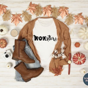 K&ouml;nnte beinhalten: Wei&szlig;es T-Shirt mit dem Text "Momster" in Schwarz mit einem Spinnennetz-Design. Das Shirt liegt auf einer Holzoberfl&auml;che mit einem braunen Cardigan, Jeans, braunen Stiefeln und Herbstdeko.