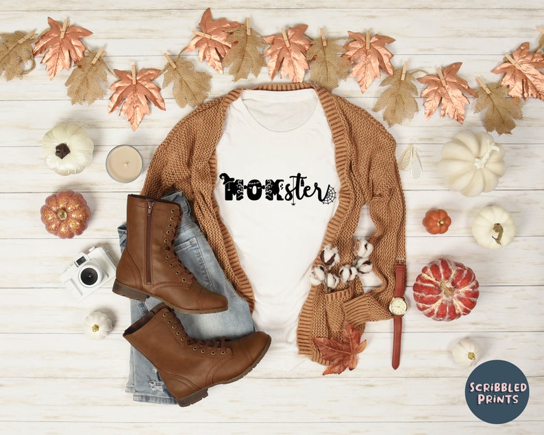 K&ouml;nnte beinhalten: Wei&szlig;es T-Shirt mit dem Text "Momster" in Schwarz mit einem Spinnennetz-Design. Das Shirt liegt auf einer Holzoberfl&auml;che mit einem braunen Cardigan, Jeans, braunen Stiefeln und Herbstdeko.