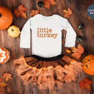 May include: White long-sleeve bodysuit with the text "little turkey" in orange. The bodysuit is paired with a tulle skirt in orange and black. The image is surrounded by fall-themed decorations, including pumpkins, leaves, and black cat ornaments.