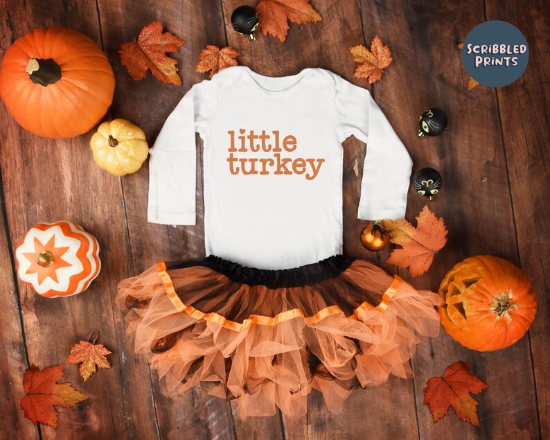 May include: White long-sleeve bodysuit with the text "little turkey" in orange. The bodysuit is paired with a tulle skirt in orange and black. The image is surrounded by fall-themed decorations, including pumpkins, leaves, and black cat ornaments.