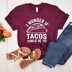 May include: A maroon t-shirt with white text that reads "I wonder if tacos think of me too". The shirt features a white graphic of a taco. The shirt is laid out on a white wooden surface with a pair of gold sandals, blue jeans, and a bouquet of pink and orange roses.