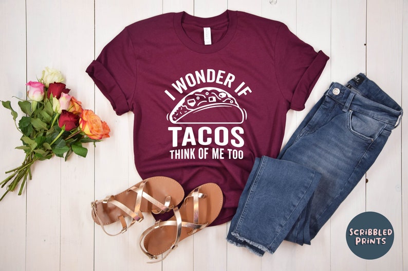 May include: A maroon t-shirt with white text that reads "I wonder if tacos think of me too". The shirt features a white graphic of a taco. The shirt is laid out on a white wooden surface with a pair of gold sandals, blue jeans, and a bouquet of pink and orange roses.