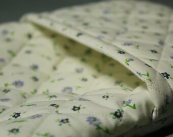 May include: A white quilted fabric with a small floral pattern in blue and green. The fabric has a pocket or flap that is folded over.