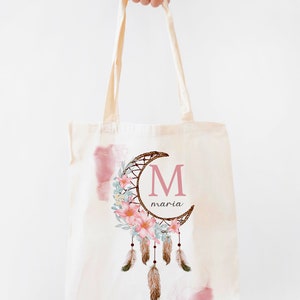 Puede incluir: Una bolsa de lona blanca con un diseño de atrapasueños rosa y marrón. El atrapasueños presenta una luna creciente con una corona de flores y plumas. La letra "M" está en el centro de la luna con el nombre "maria" escrito debajo.