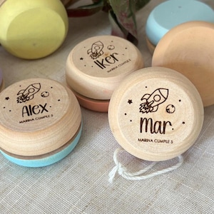 Może przedstawiać: Drewniane jo-jo z personalizowanymi imionami i grafiką rakiet. Jo-jo są w różnych kolorach, w tym jasnoniebieskim, brzoskwiniowym i żółtym. Każde jo-jo ma imię i tekst "Marina Cumple 5".