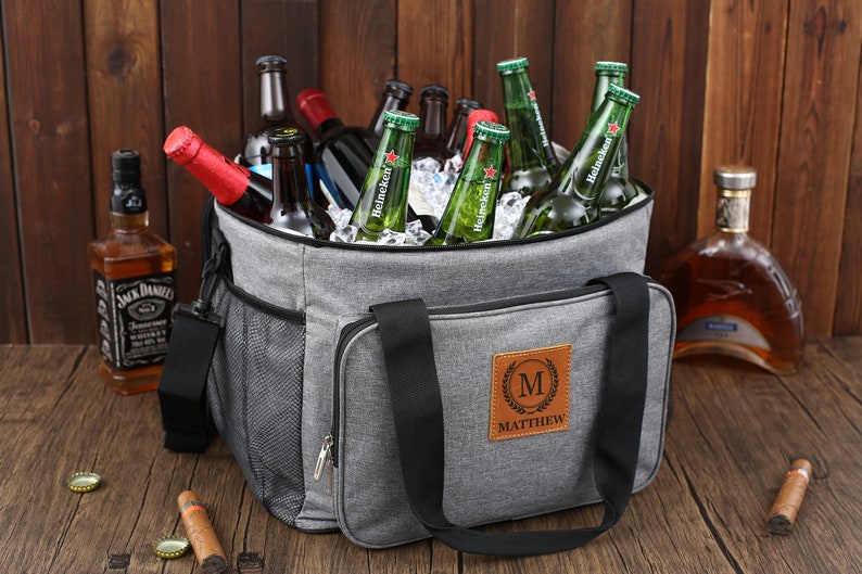 May include: A gray insulated cooler bag with a black handle and a brown leather patch that says "MATTHEW" in a gold font. The cooler bag is filled with ice and bottles of beer.