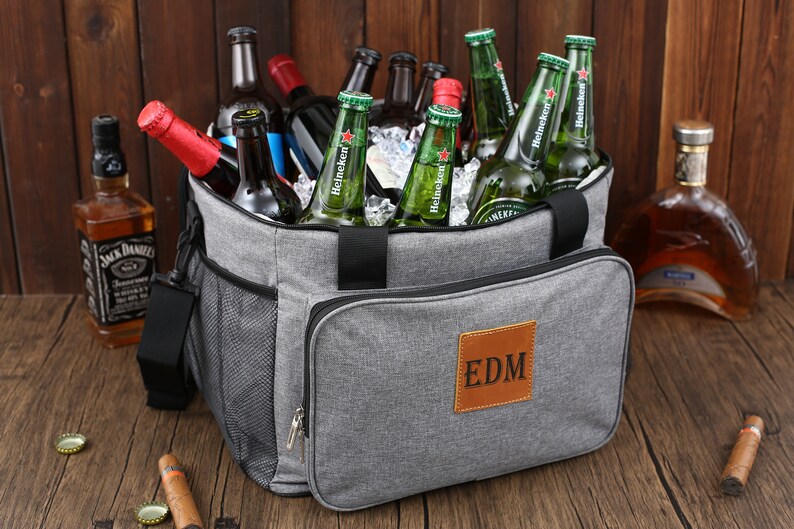 May include: A gray insulated cooler bag with a brown leather patch that says "EDM". The bag is filled with ice and bottles of beer.