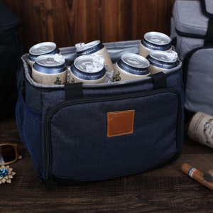 K&ouml;nnte beinhalten: Eine blaue und schwarze K&uuml;hltasche mit einem braunen Lederpatch. Die Tasche ist mit Bierdosen und Eis gef&uuml;llt. Die Tasche hat eine seitliche Netztasche und eine vordere Rei&szlig;verschlusstasche.