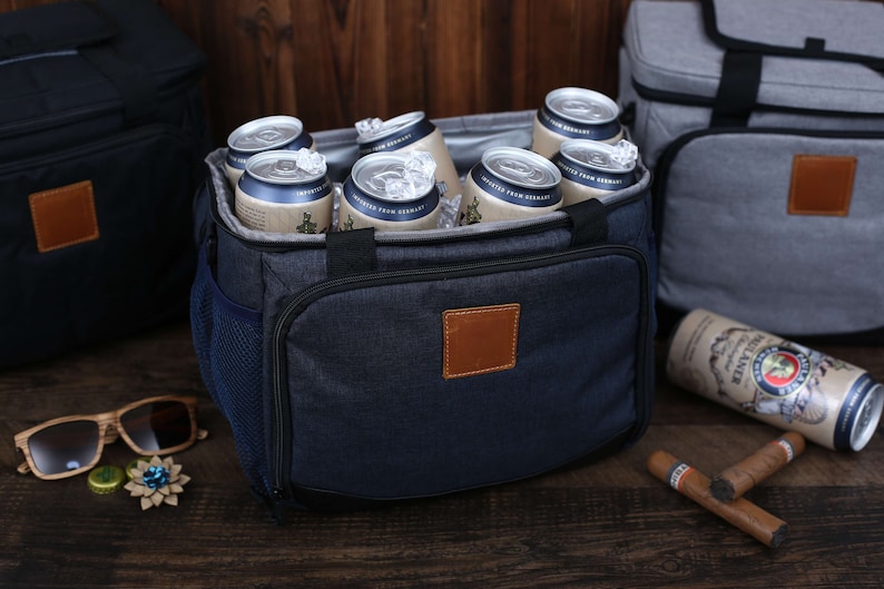 K&ouml;nnte beinhalten: Eine blaue und schwarze K&uuml;hltasche mit einem braunen Lederpatch. Die Tasche ist mit Bierdosen und Eis gef&uuml;llt. Die Tasche hat eine seitliche Netztasche und eine vordere Rei&szlig;verschlusstasche.