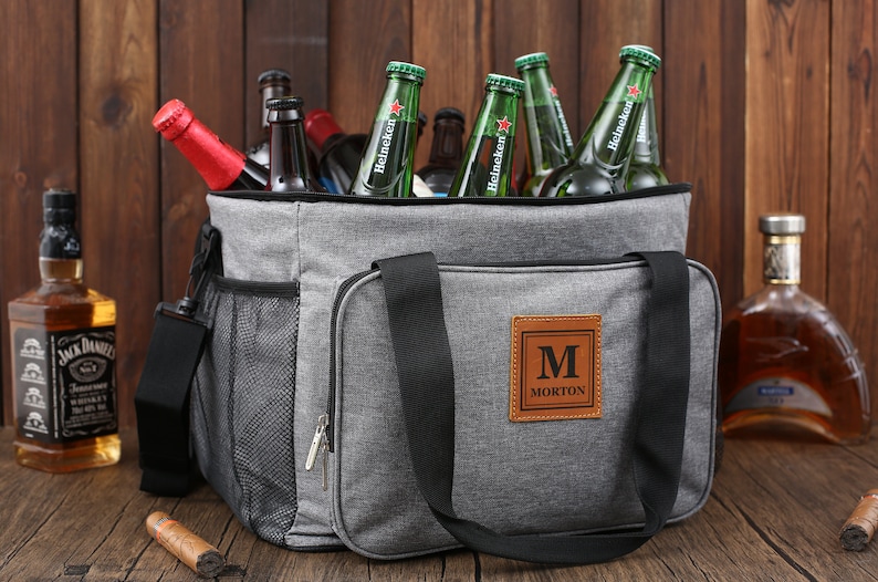 May include: A gray insulated cooler bag with a black zipper, black handles, and a brown leather patch with the letter "M" and the name "MORTON" in black text.