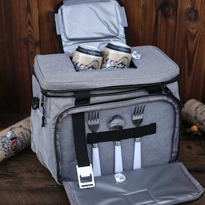 K&ouml;nnte beinhalten: Eine graue K&uuml;hltasche mit schwarzem Rei&szlig;verschluss und Griff. Die Tasche hat ein Netzfach an der Seite und ein Rei&szlig;verschlussfach an der Vorderseite. Die Tasche ist offen und enth&auml;lt vier silberne Besteckteile. Au&szlig;erdem befinden sich zwei Dosen Bier in der K&uuml;hltasche.
