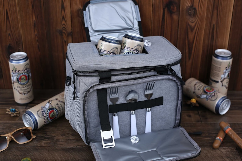 K&ouml;nnte beinhalten: Eine graue K&uuml;hltasche mit schwarzem Rei&szlig;verschluss und Griff. Die Tasche hat ein Netzfach an der Seite und ein Rei&szlig;verschlussfach an der Vorderseite. Die Tasche ist offen und enth&auml;lt vier silberne Besteckteile. Au&szlig;erdem befinden sich zwei Dosen Bier in der K&uuml;hltasche.