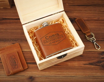 May include: A brown leather hip flask with the text "CHRIS GROOMSMEN 10-08-2021" engraved on it, inside a wooden box with a brown leather wallet with the same text engraved on it.