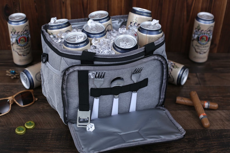 K&ouml;nnte beinhalten: Eine graue, isolierte K&uuml;hltasche mit Rei&szlig;verschluss&ouml;ffnung, ausgestattet mit einem integrierten Flaschen&ouml;ffner und einem Riemen zum Halten von Besteck. Die K&uuml;hltasche ist mit Eis und Bierdosen gef&uuml;llt.