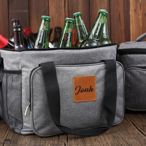 May include: Gray insulated cooler bag with a black zipper and a brown leather patch with the name "Jonh" on it. The cooler bag is filled with green glass bottles of beer.