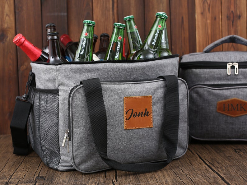 May include: Gray insulated cooler bag with a black zipper and a brown leather patch with the name "Jonh" on it. The cooler bag is filled with green glass bottles of beer.