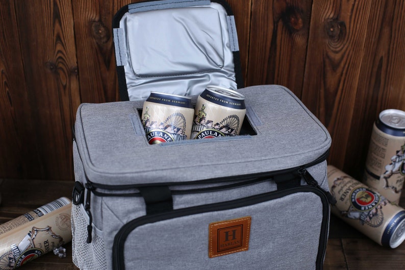 K&ouml;nnte beinhalten: Eine graue, isolierte K&uuml;hltasche mit schwarzem Rei&szlig;verschluss und Griff. Die K&uuml;hltasche hat einen braunen Lederfleck mit dem Buchstaben "H" und dem Wort "HAMMER" darauf. Zwei Dosen Bier befinden sich in der K&uuml;hltasche.
