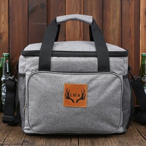 May include: A gray insulated cooler bag with black handles and a brown leather patch with the initials "LWA" and a deer antler design. The bag has a zippered pocket on the side and a mesh pocket on the front.
