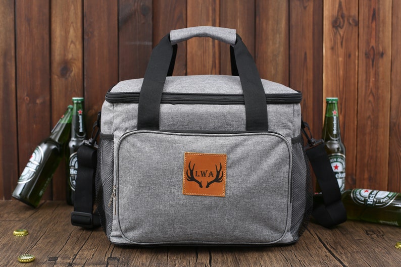 May include: A gray insulated cooler bag with black handles and a brown leather patch with the initials "LWA" and a deer antler design. The bag has a zippered pocket on the side and a mesh pocket on the front.