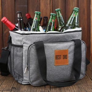 May include: A gray insulated cooler bag with black straps and a brown leather patch that says "BEST DAD". The bag is filled with green glass bottles of beer.