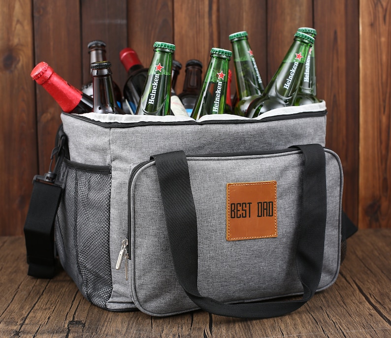 May include: A gray insulated cooler bag with black straps and a brown leather patch that says "BEST DAD". The bag is filled with green glass bottles of beer.
