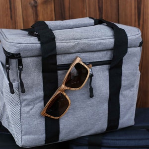 K&ouml;nnte beinhalten: Eine graue, isolierte Lunch-Tasche mit schwarzen Tr&auml;gern und einem Rei&szlig;verschluss. Ein Paar Sonnenbrillen mit Holzrahmen h&auml;ngt an der Rei&szlig;verschlusslasche.
