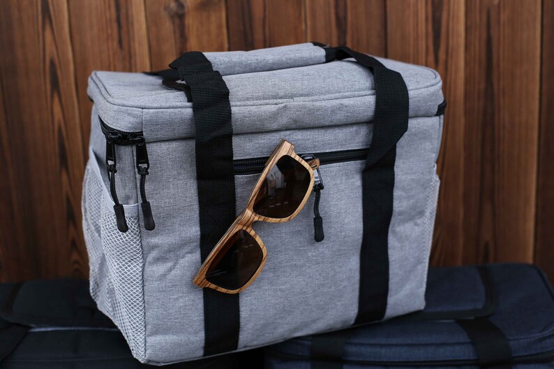 K&ouml;nnte beinhalten: Eine graue, isolierte Lunch-Tasche mit schwarzen Tr&auml;gern und einem Rei&szlig;verschluss. Ein Paar Sonnenbrillen mit Holzrahmen h&auml;ngt an der Rei&szlig;verschlusslasche.