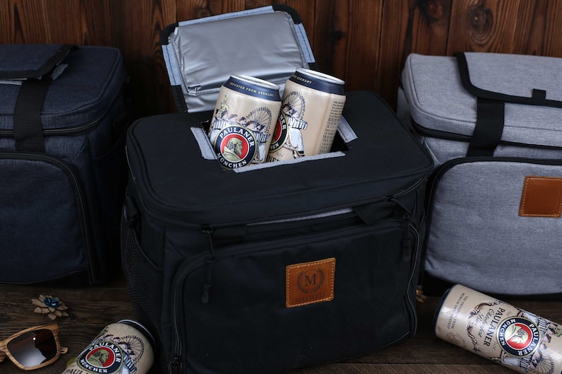 K&ouml;nnte beinhalten: Eine schwarze, isolierte K&uuml;hltasche mit einem braunen Lederflicken, auf dem ein "M" steht, und einer Rei&szlig;verschlusstasche. Die K&uuml;hltasche ist ge&ouml;ffnet und enth&auml;lt zwei Dosen Paulaner Bier. Im Hintergrund befinden sich zwei weitere K&uuml;hltaschen, eine blaue und eine graue.