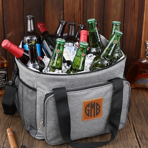 May include: A gray insulated cooler bag with black handles and a brown leather patch with the monogram "GMB". The cooler bag is filled with ice and various bottles of beer and liquor.