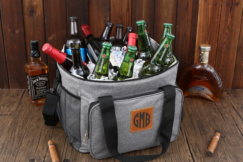 May include: A gray insulated cooler bag with black handles and a brown leather patch with the monogram "GMB". The cooler bag is filled with ice and various bottles of beer and liquor.