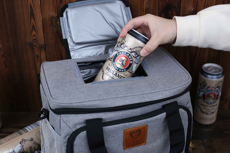 K&ouml;nnte beinhalten: Eine graue K&uuml;hltasche mit schwarzem Rei&szlig;verschluss und schwarzem Griff. Die K&uuml;hltasche hat eine Netztasche an der Seite und ein braunes Leder-Patch mit einem Logo. Eine Dose Paulaner Bier wird in die K&uuml;hltasche gelegt.