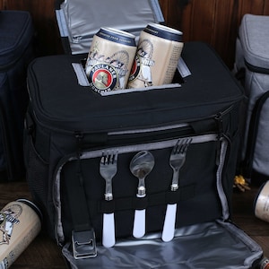 K&ouml;nnte beinhalten: Eine schwarze isolierte K&uuml;hltasche mit Rei&szlig;verschluss oben und einem Netzfach an der Seite. Die K&uuml;hltasche hat einen integrierten Besteckhalter mit drei F&auml;chern f&uuml;r Gabeln und L&ouml;ffel. Zwei Dosen Paulaner Bier sind in der K&uuml;hltasche sichtbar.