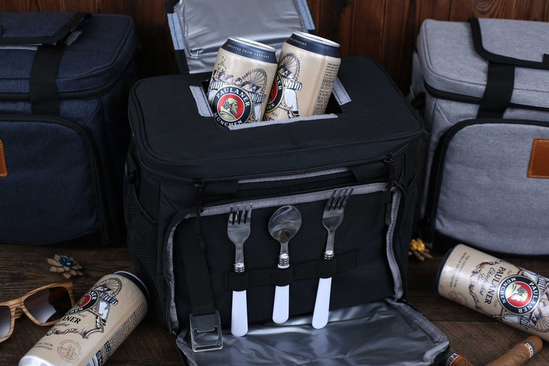 K&ouml;nnte beinhalten: Eine schwarze isolierte K&uuml;hltasche mit Rei&szlig;verschluss oben und einem Netzfach an der Seite. Die K&uuml;hltasche hat einen integrierten Besteckhalter mit drei F&auml;chern f&uuml;r Gabeln und L&ouml;ffel. Zwei Dosen Paulaner Bier sind in der K&uuml;hltasche sichtbar.