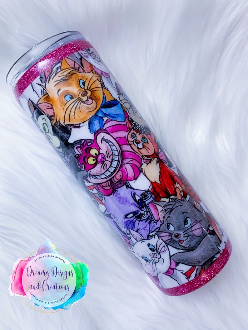 May include: A pink and white tumbler with a glitter rim features a colourful collage of cartoon cats from the Disney movie The Aristocats. The tumbler has the text "Dreamy Designs and Creations" on the side.