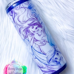 Puede incluir: Un vaso de acero inoxidable con fondo azul brillante y fondo blanco. El vaso presenta un dise&ntilde;o degradado morado y azul con un boceto de Ariel y Sebasti&aacute;n de La Sirenita. El texto "Dreamy Designs and Creations" es visible en el vaso.