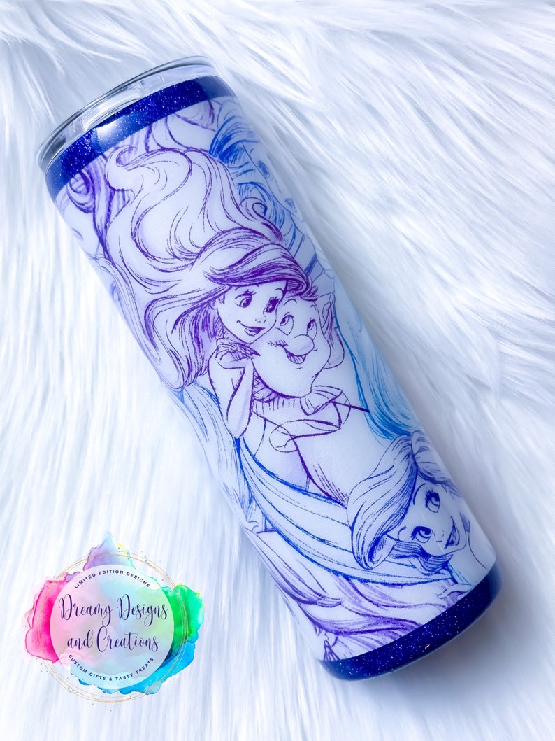 Puede incluir: Un vaso de acero inoxidable con fondo azul brillante y fondo blanco. El vaso presenta un dise&ntilde;o degradado morado y azul con un boceto de Ariel y Sebasti&aacute;n de La Sirenita. El texto "Dreamy Designs and Creations" es visible en el vaso.