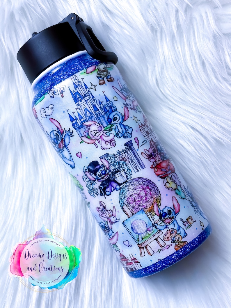 Op de afbeelding: Een blauwe en witte roestvrijstalen waterfles met een zwarte dop en handvat. De fles is versierd met een patroon van Stitch-personages uit Disney's Lilo & Stitch, samen met afbeeldingen van Assepoester's kasteel, Spaceship Earth en andere Disney-iconen. De tekst "Dreamy Designs and Creations" is op de fles gedrukt.