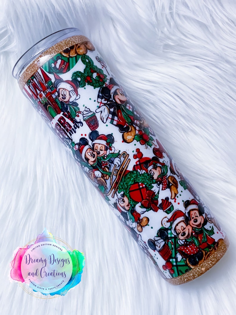 Pode incluir: Um copo branco com uma borda e base de glitter dourado. O copo &eacute; coberto com um padr&atilde;o de Natal com Mickey Mouse e Minnie Mouse em v&aacute;rias poses. O padr&atilde;o inclui xadrez vermelho e verde, azevinho e enfeites. O texto "Feliz Natal" &eacute; vis&iacute;vel no copo.