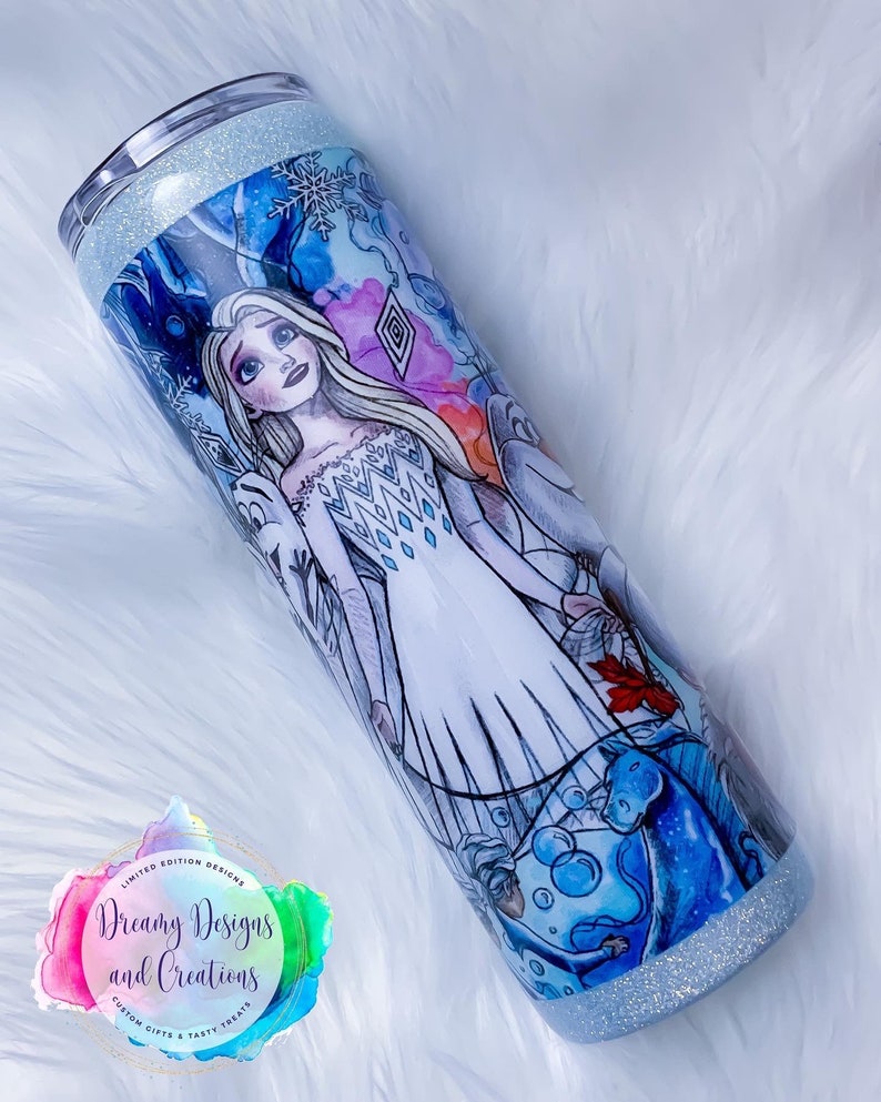 Puede incluir: Un vaso de acero inoxidable blanco y azul con purpurina, decorado con un dise&ntilde;o impreso de una mujer con el pelo largo y rubio que lleva un vestido blanco con un patr&oacute;n azul y blanco. El dise&ntilde;o tambi&eacute;n incluye copos de nieve, hielo y un caballo azul.